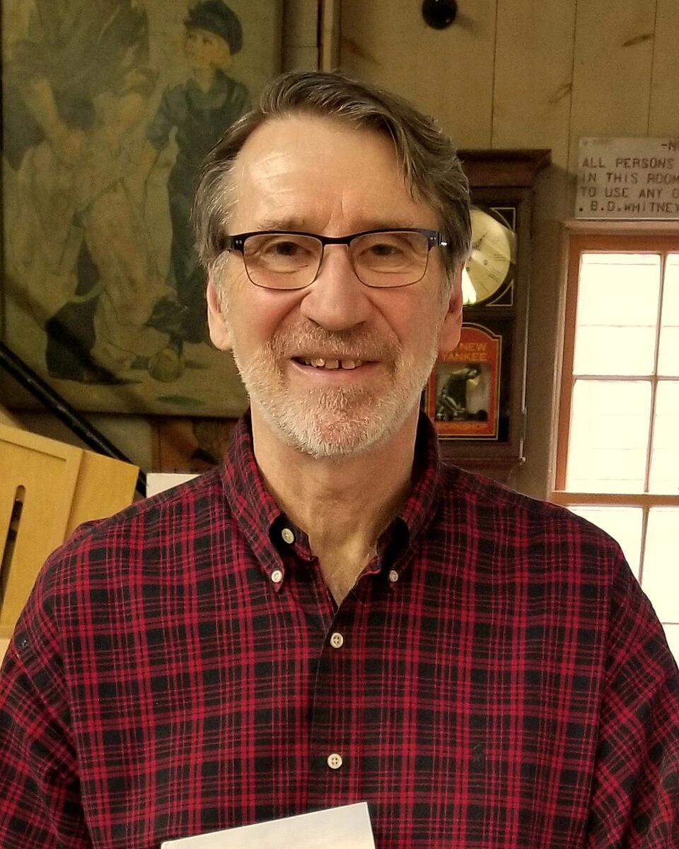 Norm Abram in a red plaid shirt and glasses standing in a workshop